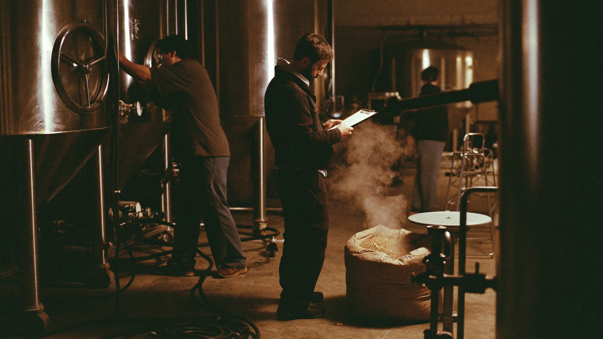 Brewers working around stainless steel tanks on a craft brewery production floor, steam drifting in warm light